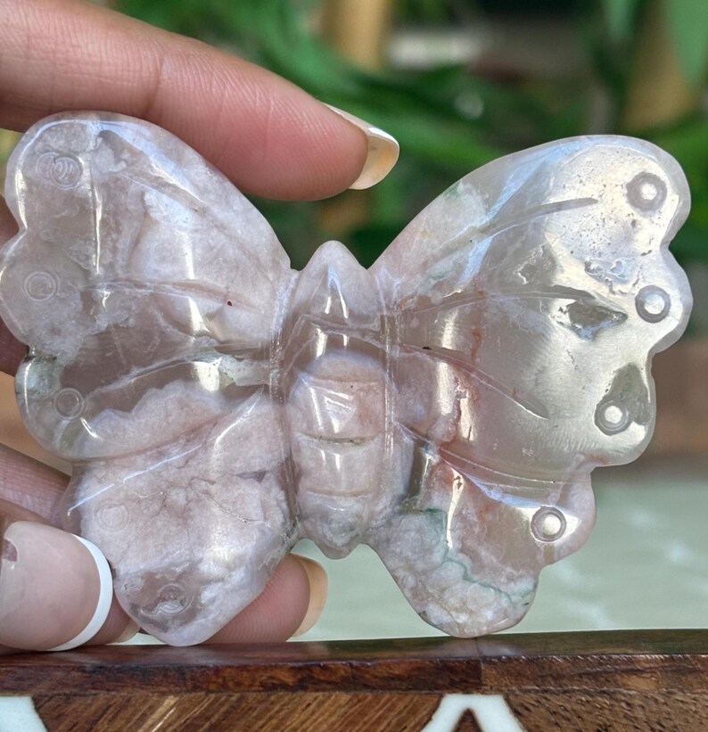 May include: A carved butterfly figurine made of translucent, light pink and white stone. The butterfly has detailed wing patterns and circular accents. The figurine is held by a hand, showcasing the intricate details and natural variations in the stone.