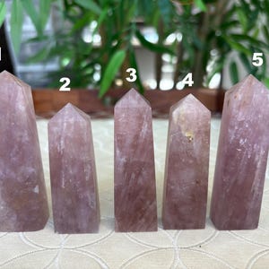 Op de afbeelding: Vijf roze kwarts kristallen obelisken van verschillende hoogtes worden getoond. Elke obelisk heeft een rechthoekige basis en een puntige top. De kristallen hebben een doorschijnend uiterlijk met subtiele kleur- en textuurvariaties.
