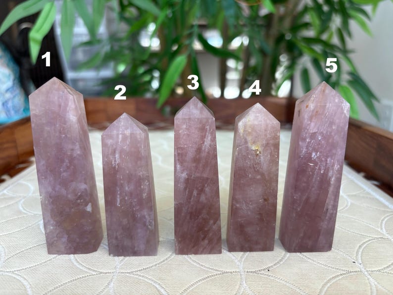 Op de afbeelding: Vijf roze kwarts kristallen obelisken van verschillende hoogtes worden getoond. Elke obelisk heeft een rechthoekige basis en een puntige top. De kristallen hebben een doorschijnend uiterlijk met subtiele kleur- en textuurvariaties.