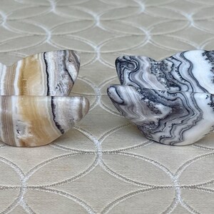 May include: Two pairs of heart-shaped stone bowls. One pair features tan, brown, and white striped patterns. The other pair has a black, gray, and white marbled design. The bowls are displayed on a neutral-toned surface.