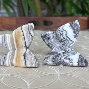 May include: Two butterfly-shaped stone sculptures. One is beige, brown, and gray, the other is black, white, and gray. The sculptures have a marbled appearance and are displayed on a patterned surface.