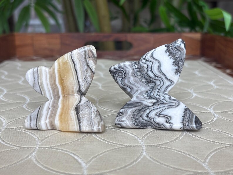 May include: Two butterfly-shaped stone sculptures. One is beige, brown, and gray, the other is black, white, and gray. The sculptures have a marbled appearance and are displayed on a patterned surface.