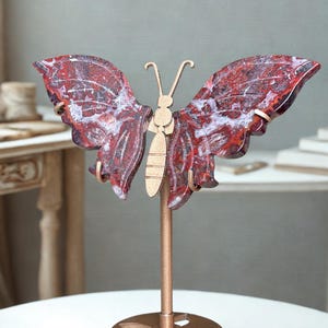 May include: A decorative butterfly sculpture featuring two polished, deep red and white marbled stone wings attached to a central gold-toned metal body. The butterfly is mounted on a slender gold-toned metal rod with a circular, cutout base.