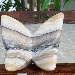 May include: A butterfly-shaped sculpture carved from layered stone, showcasing bands of beige, brown, and gray. The wings are rounded and smooth, with a polished finish. The sculpture is displayed on a wooden surface with a decorative inlay.