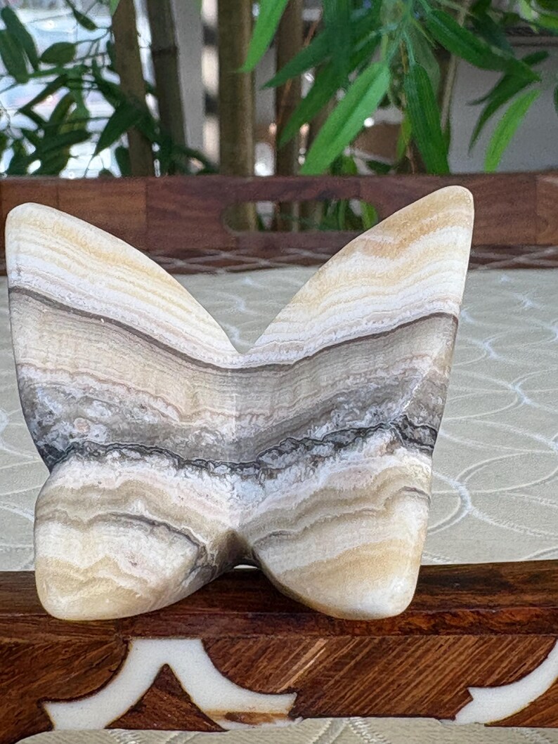 May include: A butterfly-shaped sculpture carved from layered stone, showcasing bands of beige, brown, and gray. The wings are rounded and smooth, with a polished finish. The sculpture is displayed on a wooden surface with a decorative inlay.