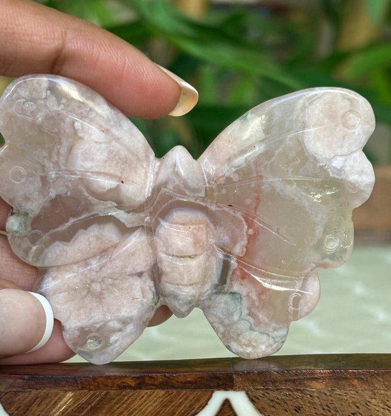 May include: A carved butterfly figurine made of a light pink and white stone. The wings have a detailed pattern with hints of green and red. The butterfly is held by a hand, showcasing the intricate details of the stone carving.