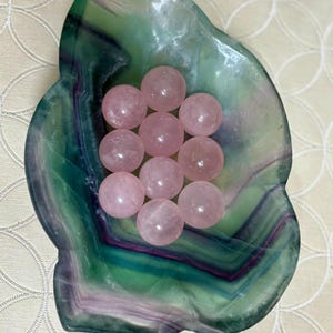 May include: A collection of ten pink rose quartz spheres nestled in a green and purple fluorite bowl. The bowl has a natural, organic shape and a polished surface. The spheres are smooth and translucent, with a soft, rosy hue. The arrangement is displayed on a cream-colored surface.