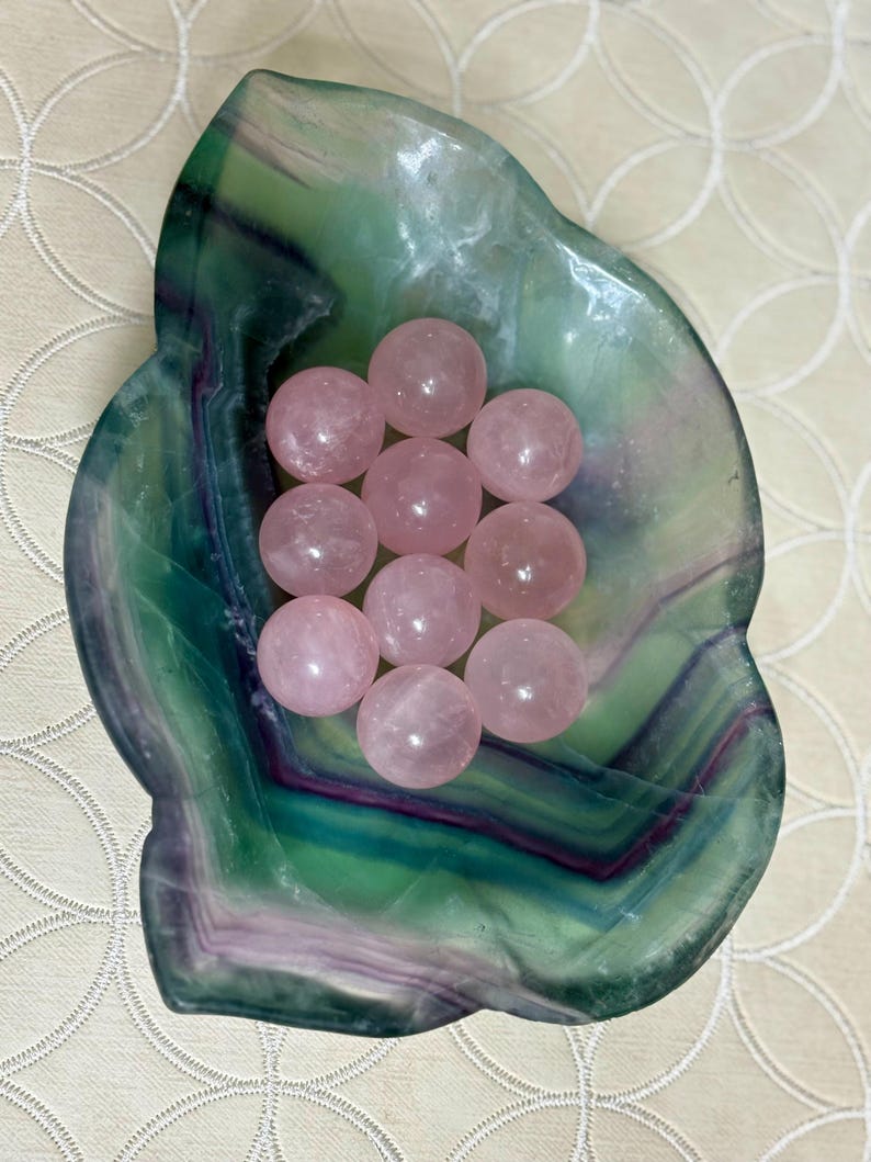 May include: A collection of ten pink rose quartz spheres nestled in a green and purple fluorite bowl. The bowl has a natural, organic shape and a polished surface. The spheres are smooth and translucent, with a soft, rosy hue. The arrangement is displayed on a cream-colored surface.
