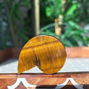 Puede incluir: Una piedra preciosa de ojo de tigre pulida, que muestra un rico color marr&oacute;n dorado con estr&iacute;as naturales. La piedra tiene una forma &uacute;nica y org&aacute;nica, que se asemeja a una hoja o l&aacute;grima estilizada. La piedra preciosa se exhibe sobre una superficie de madera decorativa.