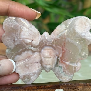 May include: A carved butterfly figurine made of a light pink and white stone. The butterfly has detailed wing patterns and a textured body. The stone has subtle red and green inclusions. The figurine is held in a hand.