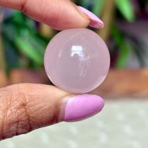 May include: A smooth, pale pink sphere, held between two fingers with pink nail polish. The sphere reflects light, suggesting a polished surface. The background is blurred, with green foliage visible.