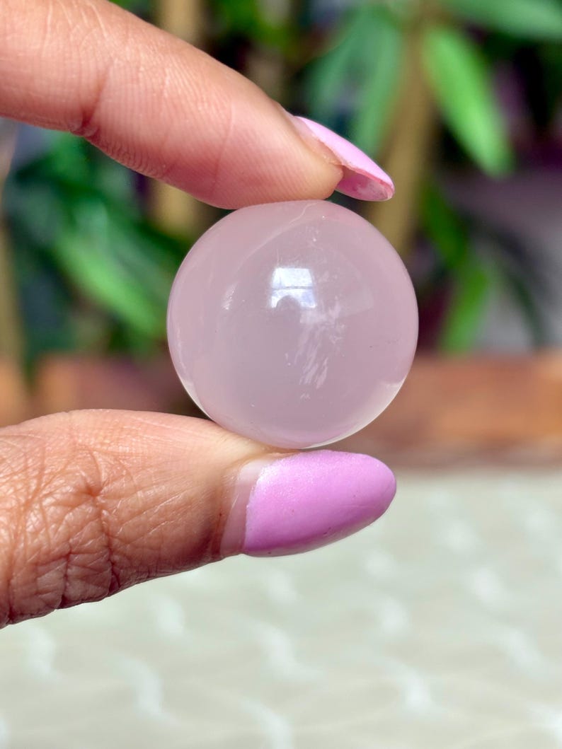 May include: A smooth, pale pink sphere, held between two fingers with pink nail polish. The sphere reflects light, suggesting a polished surface. The background is blurred, with green foliage visible.