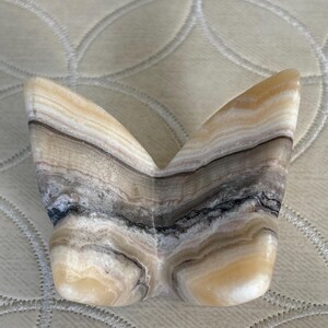 May include: A decorative butterfly sculpture carved from layered stone. The sculpture features a symmetrical design with a mix of beige, brown, gray, and black bands. The wings are spread, and the body is rounded.