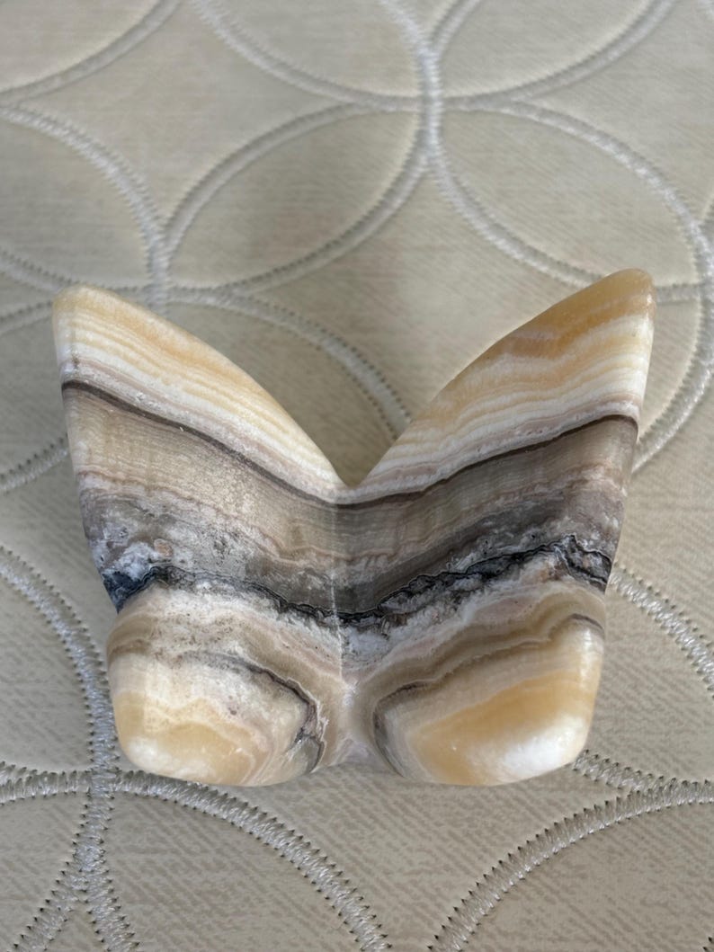 May include: A decorative butterfly sculpture carved from layered stone. The sculpture features a symmetrical design with a mix of beige, brown, gray, and black bands. The wings are spread, and the body is rounded.