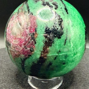 May include: A polished gemstone sphere, predominantly green with streaks of black and patches of deep red. The sphere is displayed on a clear, rounded stand, showcasing its natural patterns and colors. The gemstone is a decorative item.