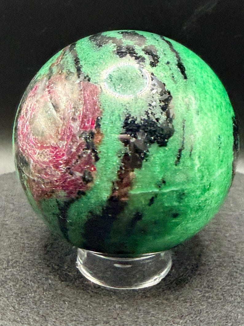 May include: A polished gemstone sphere, predominantly green with streaks of black and patches of deep red. The sphere is displayed on a clear, rounded stand, showcasing its natural patterns and colors. The gemstone is a decorative item.