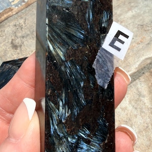 May include: A dark, rectangular crystal tower with a pointed top. The crystal has a deep brown base color with streaks of blue and white throughout. A small white sticker with the letter "E" is partially adhered to the crystal.