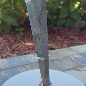 May include: A tall, six-sided crystal point with a weathered, mottled surface in shades of muted green, brown, and rust red. The crystal is held upright by a simple, circular silver-toned metal wire stand that grips the base and lower section.