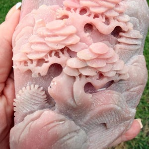 May include: A light pink stone carving featuring a detailed relief of a landscape with layered trees and rocky formations. The surface of the stone has a matte, textured finish with intricate cutouts depicting foliage and natural terrain.