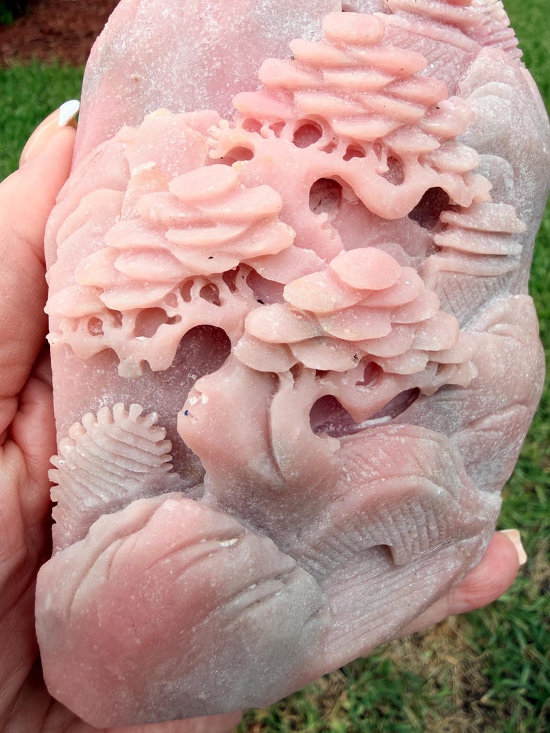 May include: A light pink stone carving featuring a detailed relief of a landscape with layered trees and rocky formations. The surface of the stone has a matte, textured finish with intricate cutouts depicting foliage and natural terrain.
