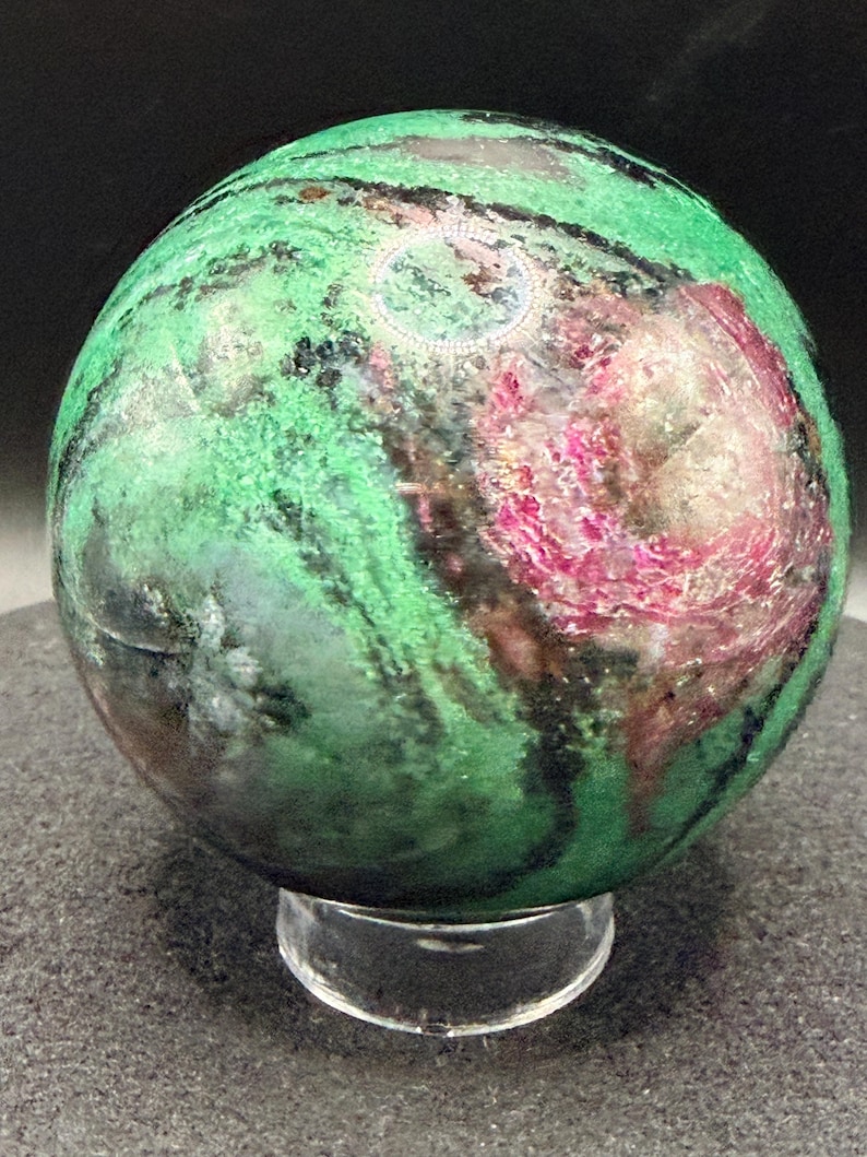 May include: A polished sphere of green and pink stone, resting on a clear acrylic stand. The stone features a mix of green, black, and pink hues, with a swirling pattern. The sphere is a decorative item.