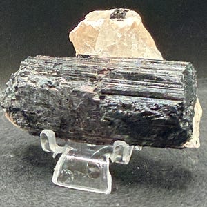 May include: A black tourmaline crystal specimen with a rough, textured surface. The crystal is displayed on a clear plastic stand, with a smaller, light-colored crystal behind it. The background is dark.
