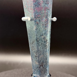 May include: A tall, faceted crystal with a blue and red mottled pattern. The crystal is mounted on a silver-colored metal stand with a circular base and two horizontal supports. The background is dark.