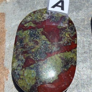 Puede incluir: Piedra ovalada con una mezcla de colores rojo, verde y negro. La piedra tiene una superficie pulida y un aspecto natural. Una peque&ntilde;a etiqueta blanca con la letra "A" est&aacute; adherida a la piedra.