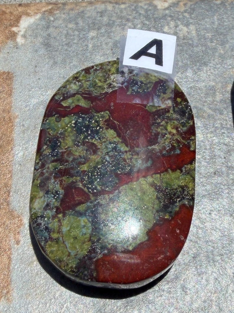 Puede incluir: Piedra ovalada con una mezcla de colores rojo, verde y negro. La piedra tiene una superficie pulida y un aspecto natural. Una peque&ntilde;a etiqueta blanca con la letra "A" est&aacute; adherida a la piedra.