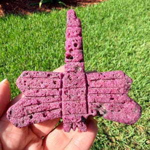 May include: A vibrant magenta dragonfly-shaped carving with a porous, textured surface. The object features a long, segmented body and two pairs of wide, horizontal wings with linear grooves carved into the surface, showcasing a rough, stone-like finish.