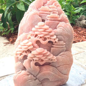 May include: A pink stone sculpture featuring an intricate relief carving of a landscape with tiered pagodas, a curved bridge, and a detailed bonsai tree. The surface of the stone has a matte texture and displays natural variations in the pink color.
