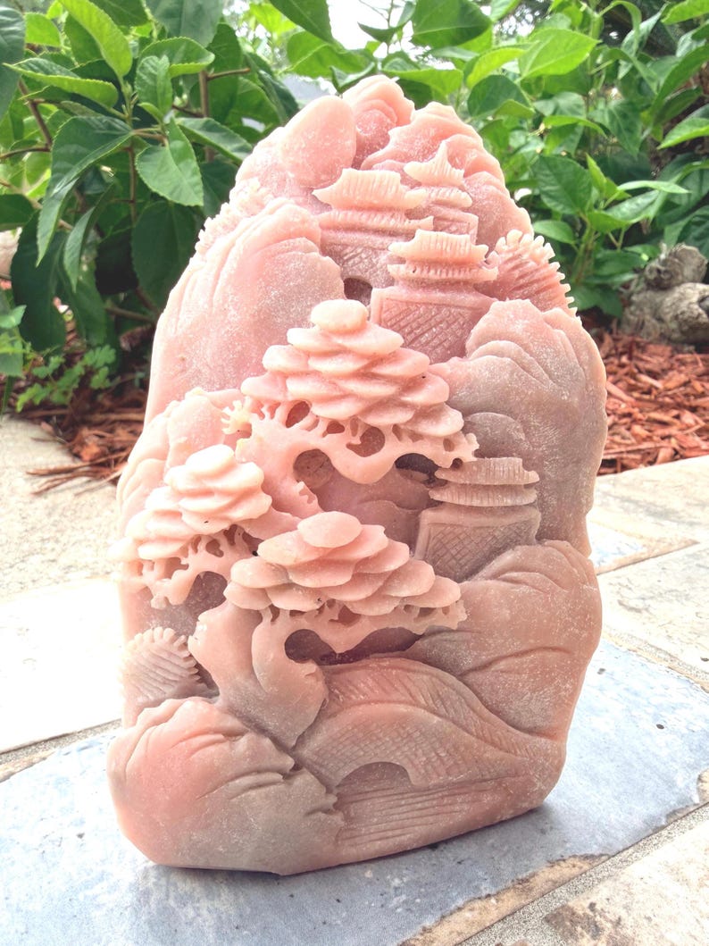 May include: A pink stone sculpture featuring an intricate relief carving of a landscape with tiered pagodas, a curved bridge, and a detailed bonsai tree. The surface of the stone has a matte texture and displays natural variations in the pink color.
