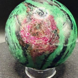 May include: A polished sphere of ruby in zoisite, showcasing a vibrant mix of green, black, and pink hues. The gemstone is displayed on a clear, rounded stand, highlighting its natural patterns and textures. This decorative sphere is a unique addition to any collection.