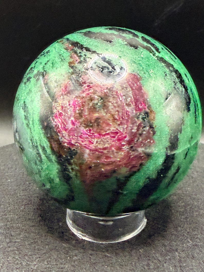 May include: A polished sphere of ruby in zoisite, showcasing a vibrant mix of green, black, and pink hues. The gemstone is displayed on a clear, rounded stand, highlighting its natural patterns and textures. This decorative sphere is a unique addition to any collection.