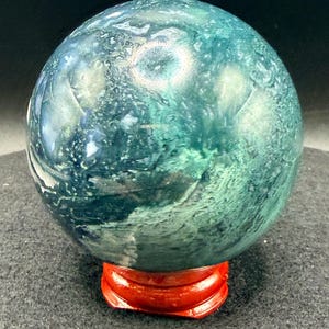 Puede incluir: Una esfera de piedra preciosa pulida, en tonos turquesa y verde, que descansa sobre un peque&ntilde;o soporte de madera rojo. El orbe tiene un aspecto veteado con patrones arremolinados. El soporte es de un rojo brillante.