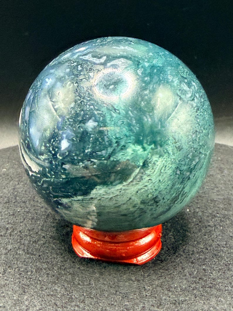 Puede incluir: Una esfera de piedra preciosa pulida, en tonos turquesa y verde, que descansa sobre un peque&ntilde;o soporte de madera rojo. El orbe tiene un aspecto veteado con patrones arremolinados. El soporte es de un rojo brillante.