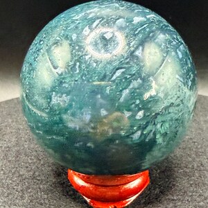 Puede incluir: Una gema esf&eacute;rica pulida, en tonos verde azulado y verde, que descansa sobre un peque&ntilde;o soporte de madera rojo. La superficie del orbe muestra un patr&oacute;n arremolinado de colores y texturas. El soporte tiene forma de media luna curvada.