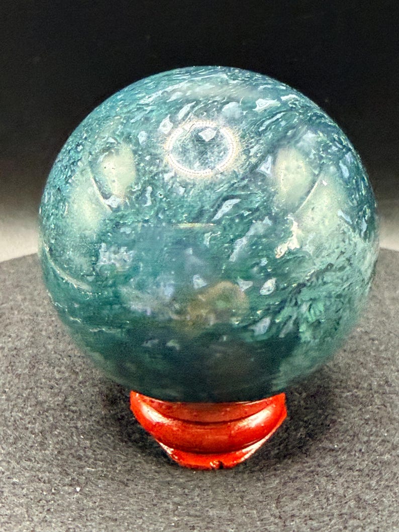 Puede incluir: Una gema esf&eacute;rica pulida, en tonos verde azulado y verde, que descansa sobre un peque&ntilde;o soporte de madera rojo. La superficie del orbe muestra un patr&oacute;n arremolinado de colores y texturas. El soporte tiene forma de media luna curvada.
