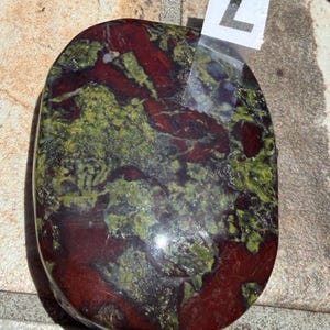 Puede incluir: Una piedra pulida de forma ovalada, con una mezcla de tonos rojo intenso y verde. La piedra tiene un acabado brillante y un aspecto natural veteado. Una peque&ntilde;a etiqueta blanca con el n&uacute;mero "7" est&aacute; adherida.