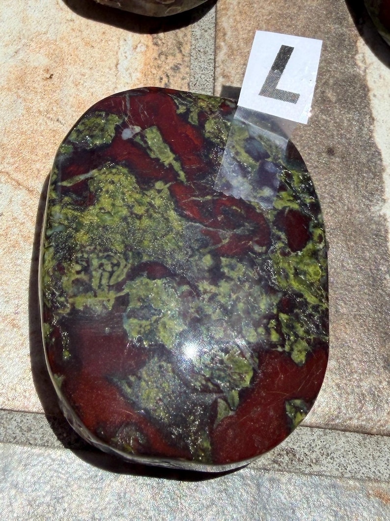Puede incluir: Una piedra pulida de forma ovalada, con una mezcla de tonos rojo intenso y verde. La piedra tiene un acabado brillante y un aspecto natural veteado. Una peque&ntilde;a etiqueta blanca con el n&uacute;mero "7" est&aacute; adherida.
