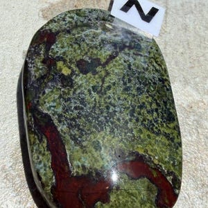 Puede incluir: Una piedra pulida de forma ovalada con un patr&oacute;n moteado de verde y rojo oscuro. La piedra tiene una superficie lisa y un aspecto natural y org&aacute;nico. Una peque&ntilde;a etiqueta blanca con la letra "N" est&aacute; adherida en la parte superior.