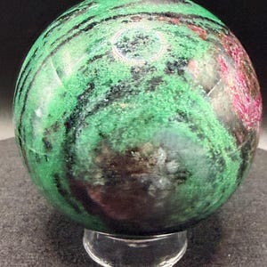 May include: A polished sphere of green zoisite with black and red inclusions, resting on a clear acrylic stand. The gemstone features a marbled appearance with varying shades of green and dark streaks. The sphere is a decorative item.
