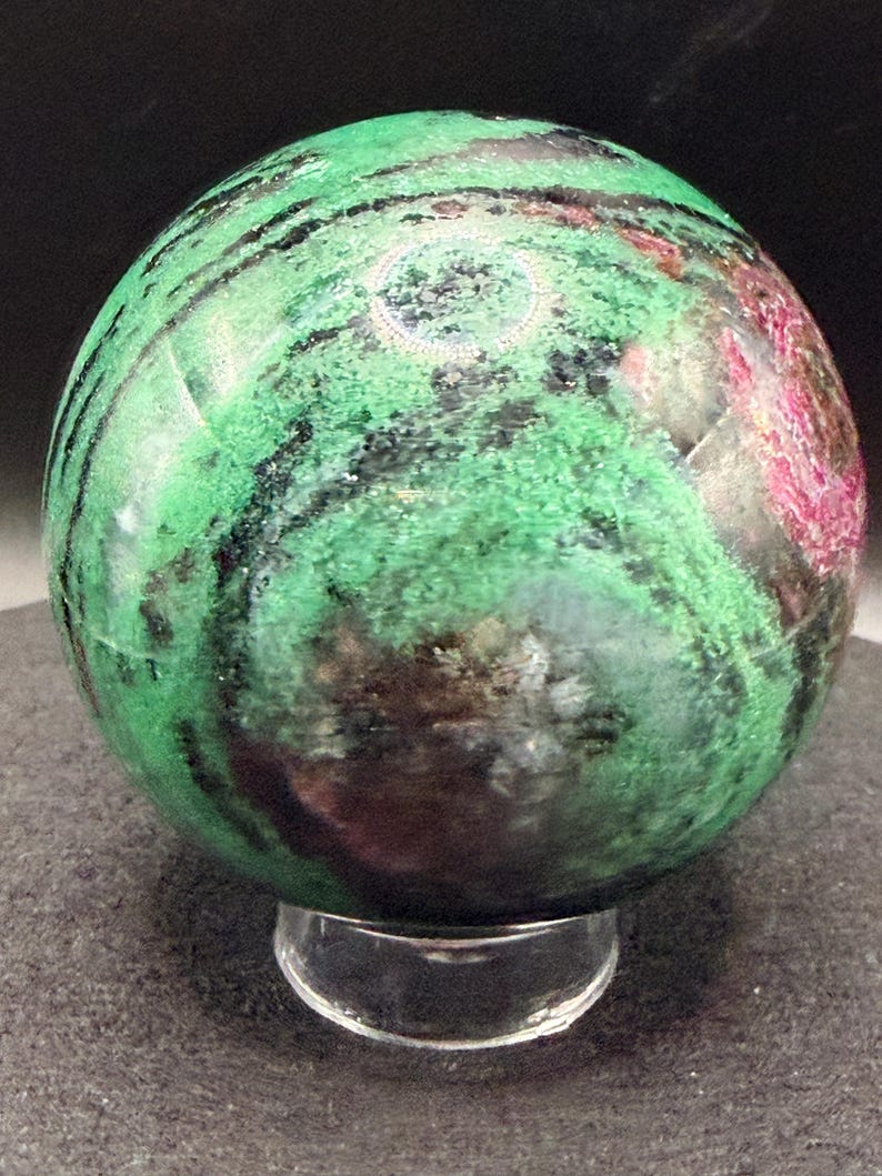 May include: A polished sphere of green zoisite with black and red inclusions, resting on a clear acrylic stand. The gemstone features a marbled appearance with varying shades of green and dark streaks. The sphere is a decorative item.