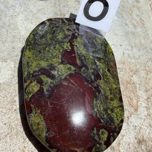 Puede incluir: Una piedra pulida de forma ovalada, con una mezcla de tonos rojo intenso y verde. La piedra tiene una superficie lisa y brillante, con un patr&oacute;n natural y org&aacute;nico. Una peque&ntilde;a etiqueta blanca con el n&uacute;mero "0" est&aacute; adjunta.