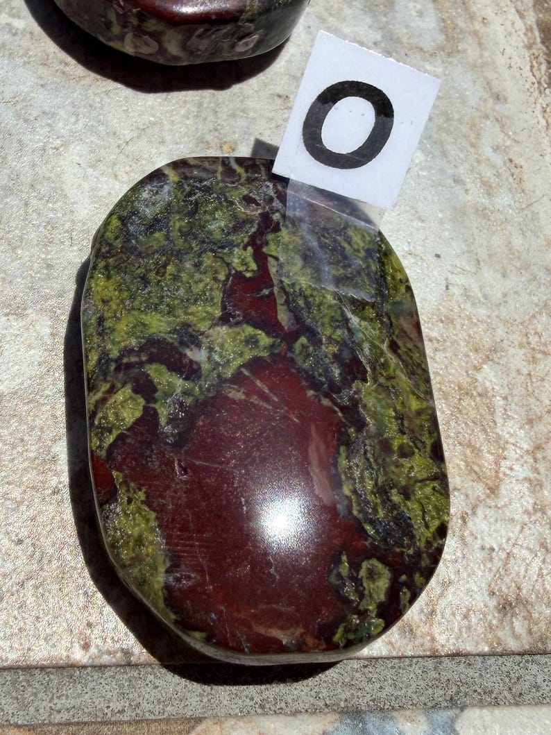 Puede incluir: Una piedra pulida de forma ovalada, con una mezcla de tonos rojo intenso y verde. La piedra tiene una superficie lisa y brillante, con un patr&oacute;n natural y org&aacute;nico. Una peque&ntilde;a etiqueta blanca con el n&uacute;mero "0" est&aacute; adjunta.