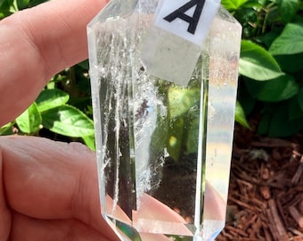 May include: A clear, double-terminated quartz crystal point featuring a white rectangular label with the black letter A printed on it. The crystal has a hexagonal shape with visible internal fractures and inclusions throughout its structure.