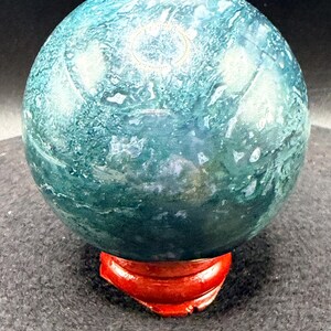 Puede incluir: Una esfera de piedra preciosa pulida con un patr&oacute;n arremolinado de color verde azulado y blanco. Se encuentra sobre un peque&ntilde;o soporte de madera rojo. La superficie de la piedra preciosa tiene un aspecto veteado, con variaciones de color.