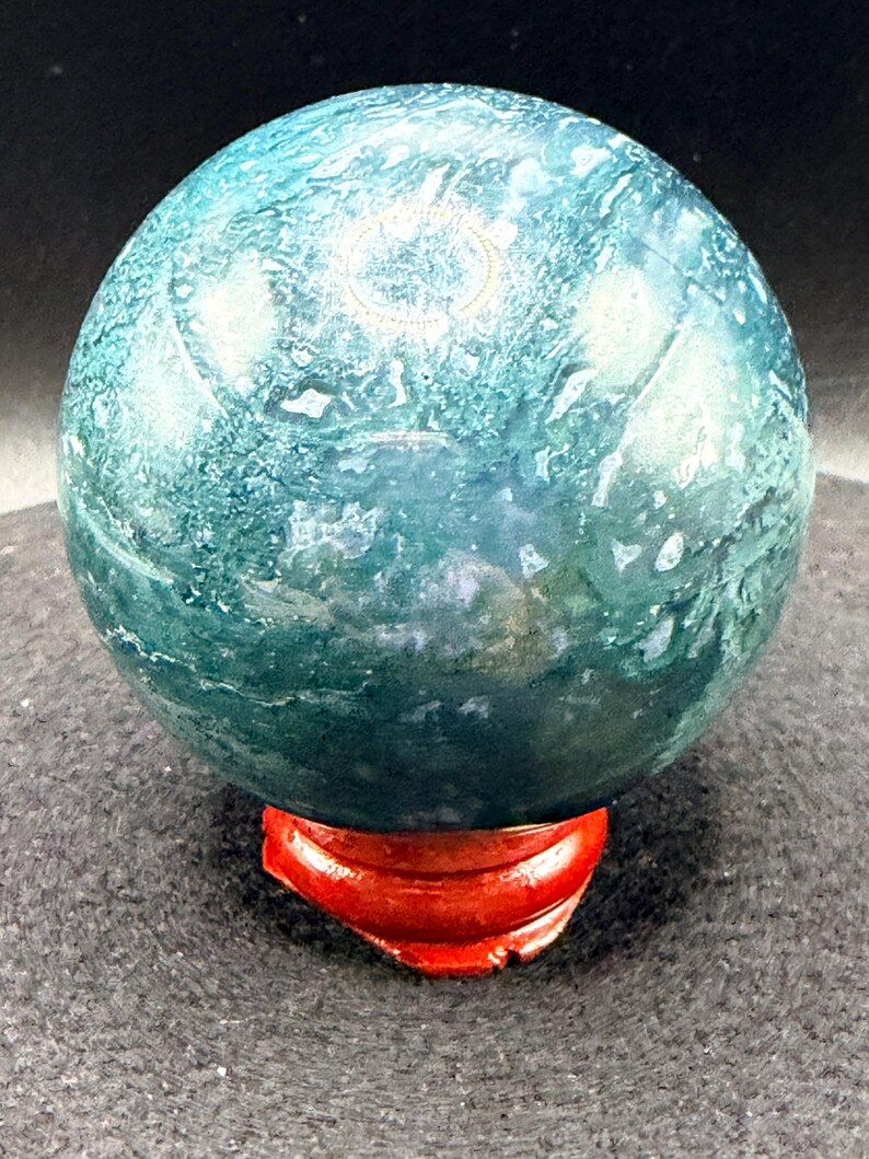 Puede incluir: Una esfera de piedra preciosa pulida con un patr&oacute;n arremolinado de color verde azulado y blanco. Se encuentra sobre un peque&ntilde;o soporte de madera rojo. La superficie de la piedra preciosa tiene un aspecto veteado, con variaciones de color.