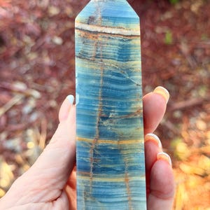 May include: A polished blue onyx obelisk with horizontal bands of color, including shades of blue, brown, and gold. The gemstone is held in a hand, showcasing its rectangular shape and pointed top. The obelisk is a decorative item.