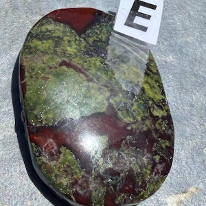 Puede incluir: Una piedra pulida de forma ovalada con una mezcla de tonos rojo oscuro y verde. La piedra tiene un acabado brillante y un aspecto natural y org&aacute;nico. Una peque&ntilde;a etiqueta blanca con la letra "E" est&aacute; adherida.